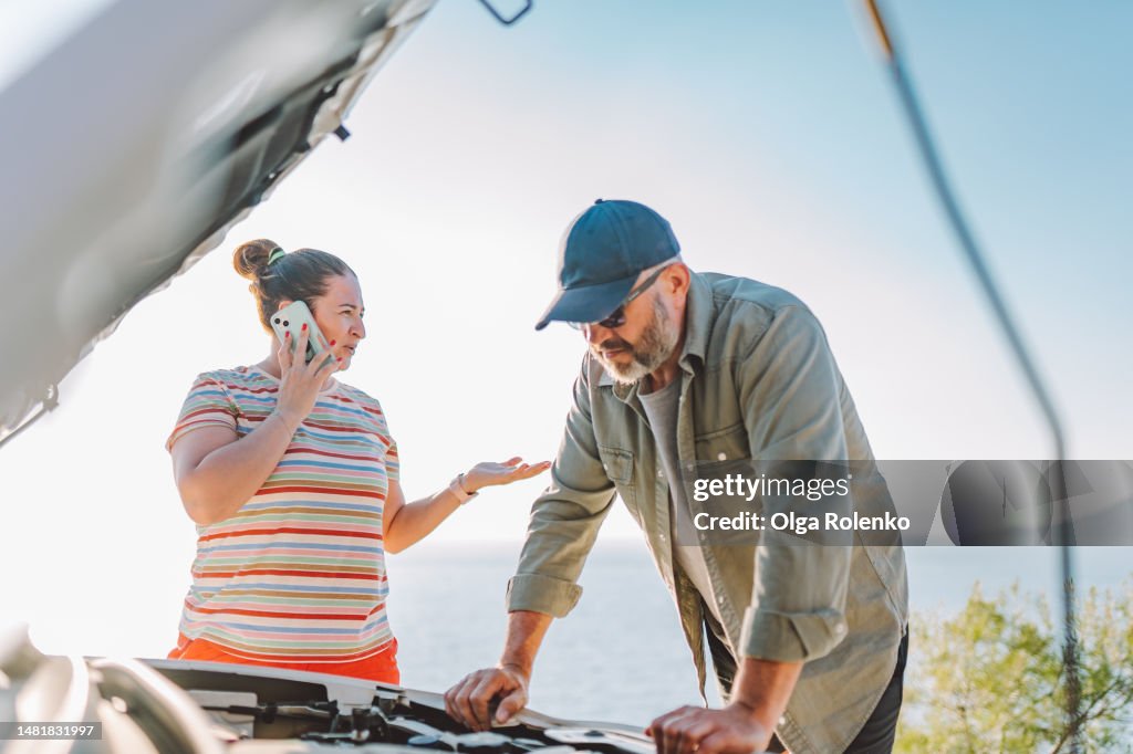 Call for help, stranded in mountains with broken car. Worried man fixing car with opened hood while woman talking on phone