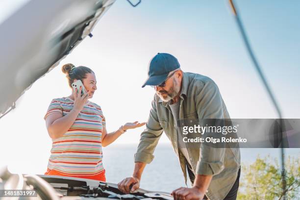 call for help, stranded in mountains with broken car. worried man fixing car with opened hood while woman talking on phone - bad appearance stock pictures, royalty-free photos & images