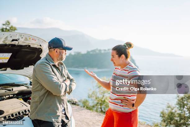 abusive relationship leads to car breakdown in mountain and sea area. woman arguing with man near opened car hood in wild mountain and sea area - vehicle breakdown - fotografias e filmes do acervo