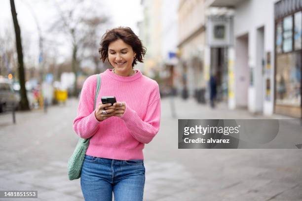 ritratto di giovane donna sorridente che usa il telefono cellulare mentre cammina all'aperto sulla strada della città - rosa germanica foto e immagini stock