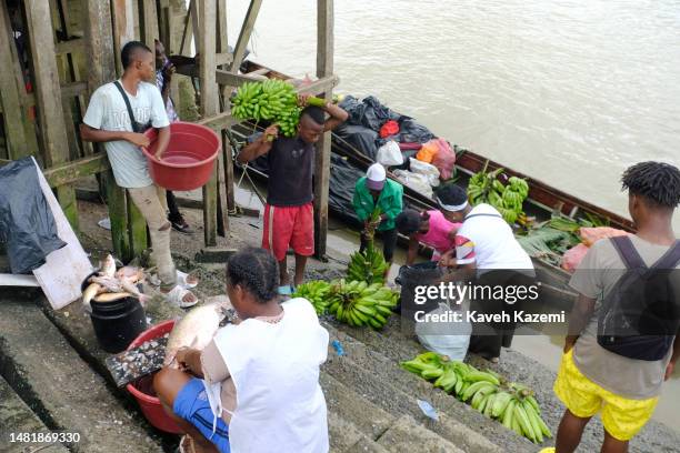 Colombian woman cleans a fish while engine powered transportation canoes bring Plantain or Platano and other tropical fruits from surrounding towns...