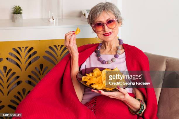 a nice stylish elderly lady in a pink dress and a large scarf is draped over her shoulders is lying on a couch in a modern living room and eating potato chips from a plate. - slim stock pictures, royalty-free photos & images
