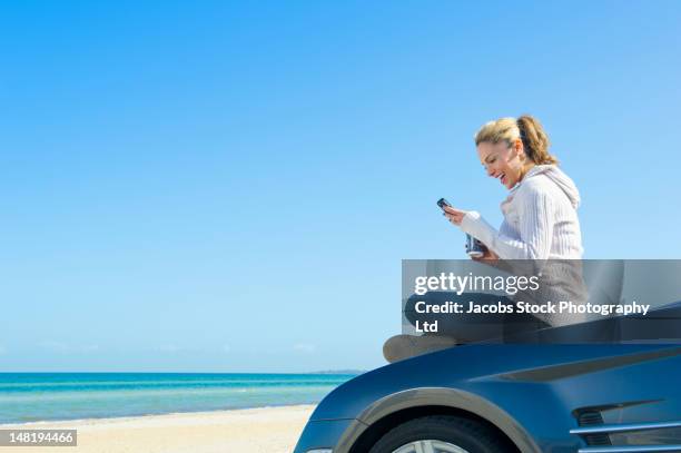 hispanic woman sitting on car at beach using cell phone - drinking soda in car stock pictures, royalty-free photos & images