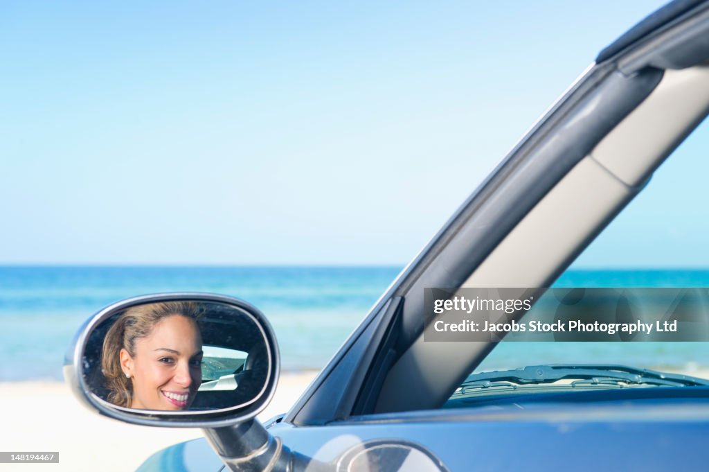 Hispanic woman on beach looking into car's rearview mirror