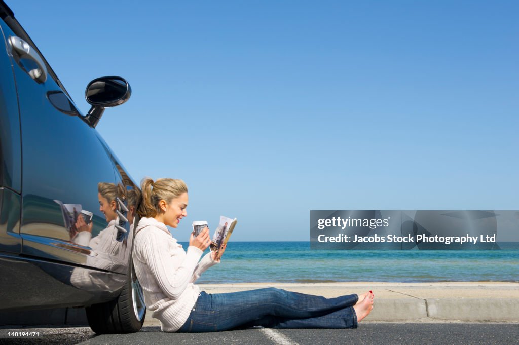 Hispanic woman leaning against car drinking coffee and reading magazine