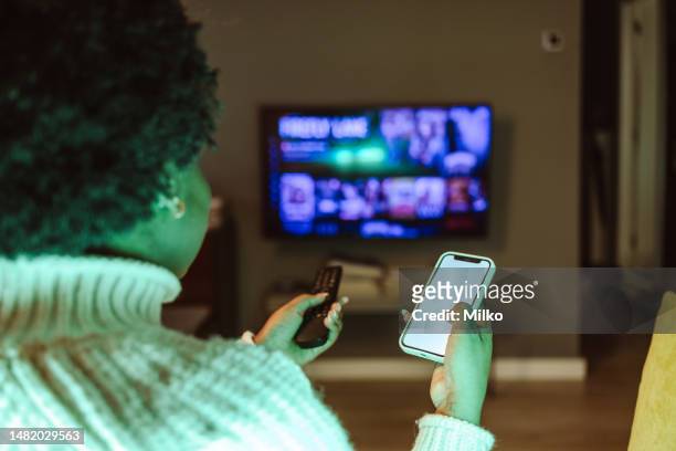 young woman holding smart phone and tv remote control - glee tv program stockfoto's en -beelden