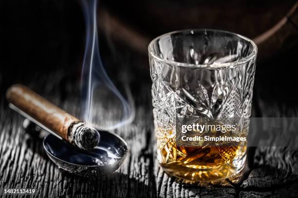 a glass of whiskey or bourbon with a burning cigar and an old wooden barrel on an old rustic table. - cognac stockfoto's en -beelden
