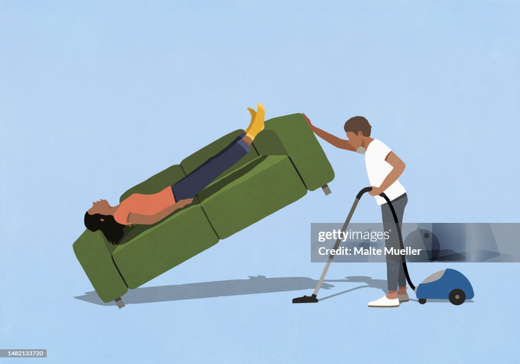 Husband vacuuming under wife sleeping on sofa at home