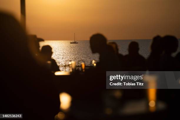 silhouetted people drinking beer on ocean waterfront patio at sunset - bar de plage photos et images de collection