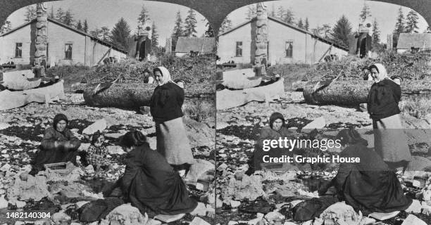 Stereoscopic image showing Haida women, one with a baby in a papoose on her back, and a small child around the laundry pool, with a totem pole in the...