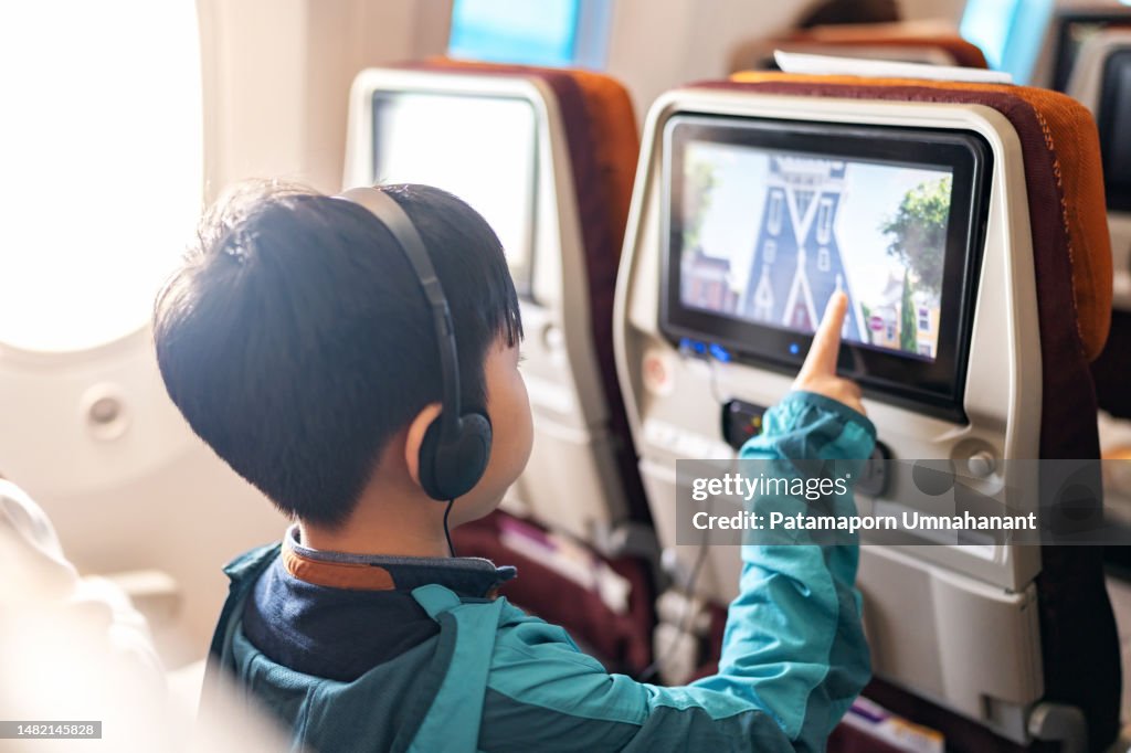 In-flight technology creates enjoyment during the journey. Back view of Asia boy wearing headphone and jacket sitting on the plane touching screen of digital tablet to choose cartoon during flight.