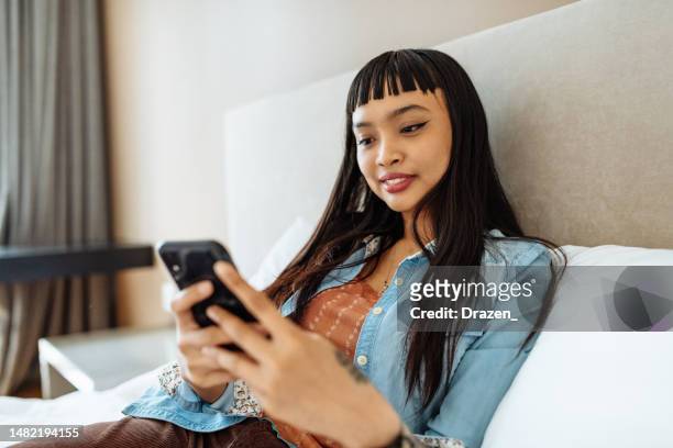 mixed race asian woman relaxing in bed and using smart phone - filipino culture stock pictures, royalty-free photos & images