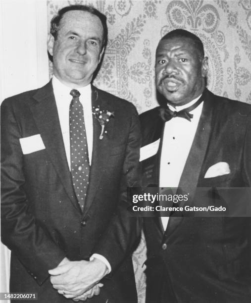 Mid shot of two men with name tags, one wearing a polka tie and a boutonniere and the other wearing a tuxedo, facing the camera with slightly...