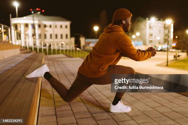 a smiling african-american man making bulgarian lunges using a bench at night in the city with the light of lanterns in the background - gluteos fotografías e imágenes de stock