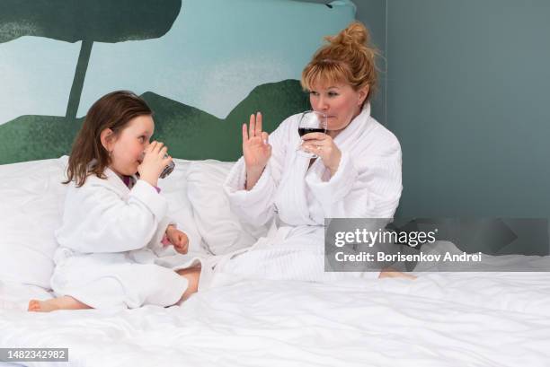 a young woman and a girl of 7 years old in dressing gowns sit on a large bed and relax with a glass of wine and a glass of juice. - lebanon wine stock pictures, royalty-free photos & images