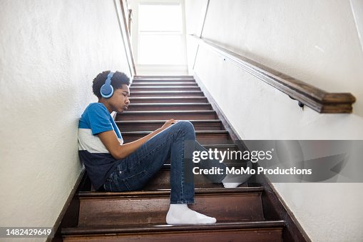 Teenage boy using wireless earphones and smartphone on staircase at home Teenage boy using wireless earphones and smartphone on staircase at home