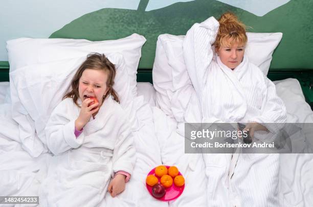 a young woman and a girl of 7 years old in home dressing gowns lie on a large bed and relax with a glass of wine, the child eats fruit. - lebanon wine stock pictures, royalty-free photos & images