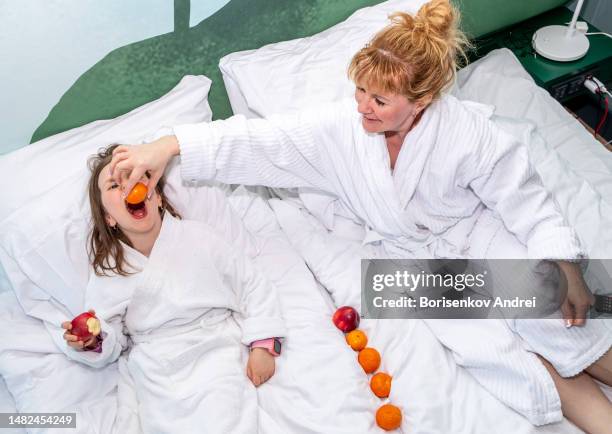 a young woman and a girl of 7 years old in home dressing gowns lie on a large bed and relax with a glass of wine, the child eats fruit. - lebanon wine stock pictures, royalty-free photos & images
