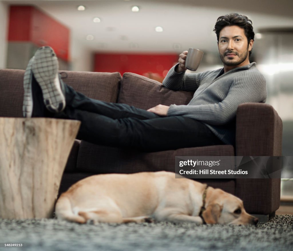 Hispanic man drinking coffee on sofa with dog nearby