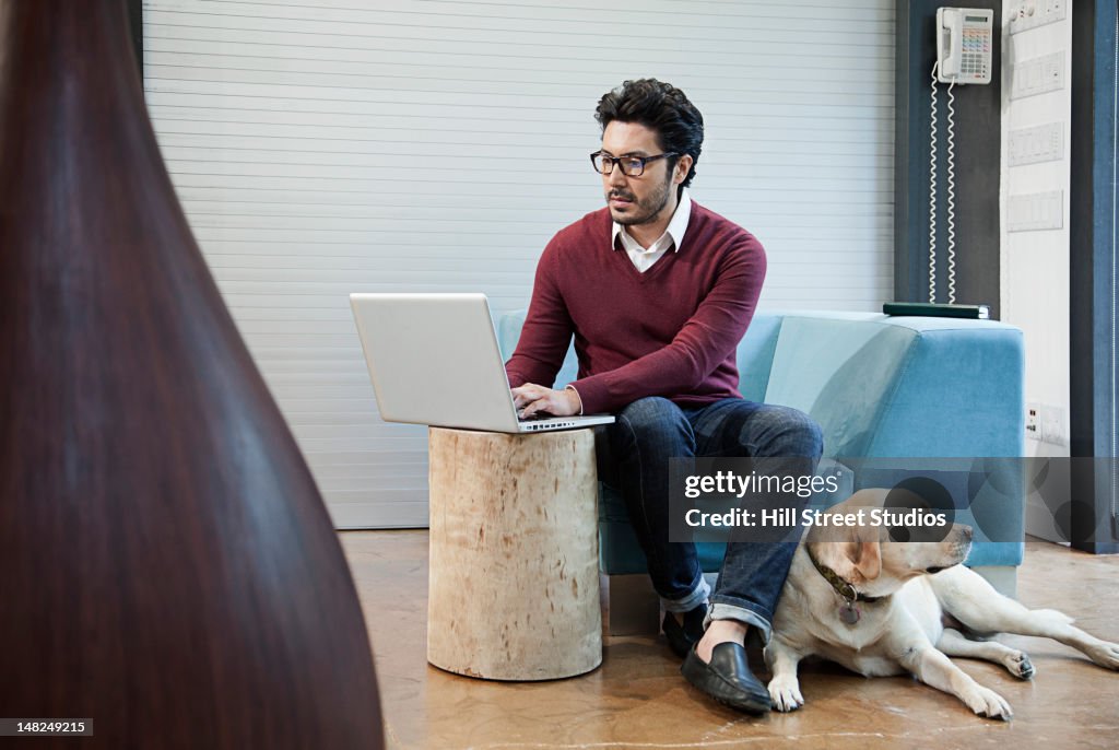 Hispanic man typing on laptop with dog by his side