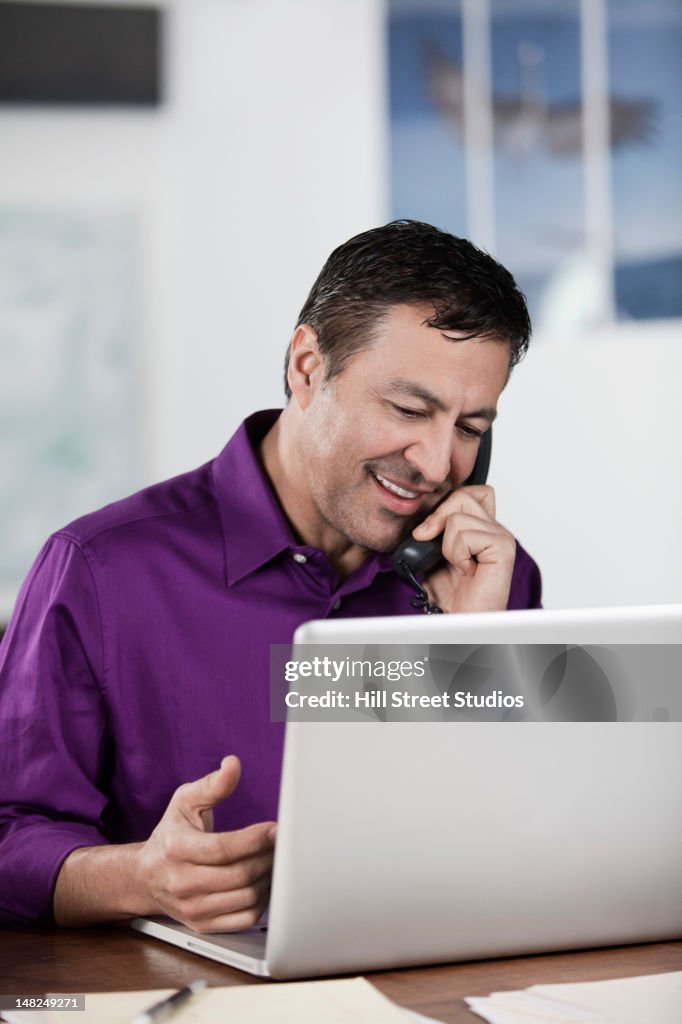 Hispanic businessman using laptop and talking on telephone