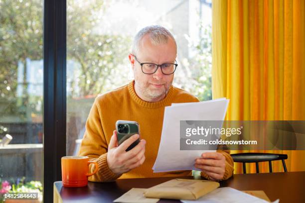 mittlerer erwachsener mann, der rechnungen mit seinem smartphone zu hause überprüft und bezahlt - high income stock-fotos und bilder