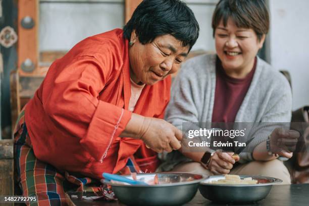 asian chinese female tourist experiential travel cutting potato in bhutanese farmhouse preparing food for dinner with bhutanese senior woman - experiential vacations stock pictures, royalty-free photos & images