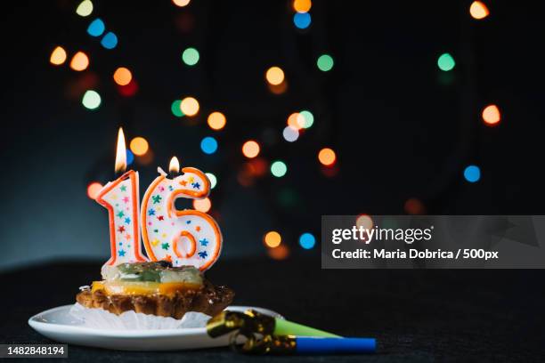 number 16 birthday lighted candle slice tart with party horn blower against illuminated bokeh backdr,romania - birthday candle number stock pictures, royalty-free photos & images