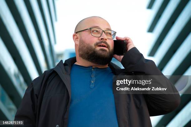 a confident young handsome man in a white shirt talks on a mobile phone. an aspiring successful businessman or manager outdoors, wearing sunglasses, looks haughtily into the camera. there is a modern car nearby. - best sunglasses for bald men fotografías e imágenes de stock