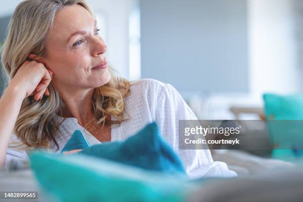 reife frau, die sich auf dem sofa entspannt. sie sieht zufrieden und nachdenklich aus - menopause stock-fotos und bilder