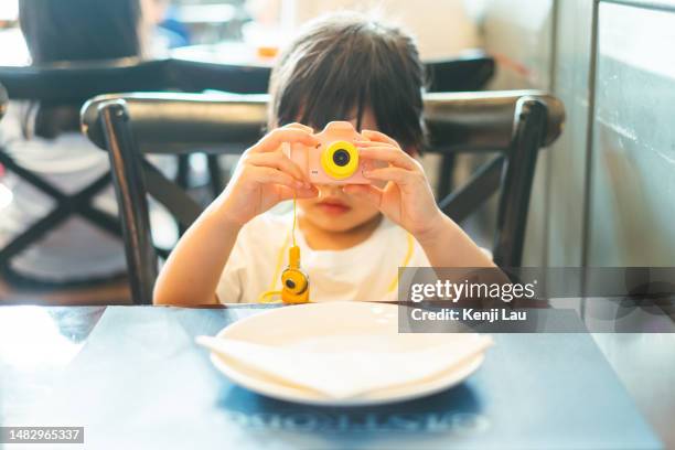 portrait of a little asian girl playing a toy camera while waiting for food with plain plate in restaurant. having a fun time dining out with her family. family concept. family eating out lifestyle - toy camera stock pictures, royalty-free photos & images