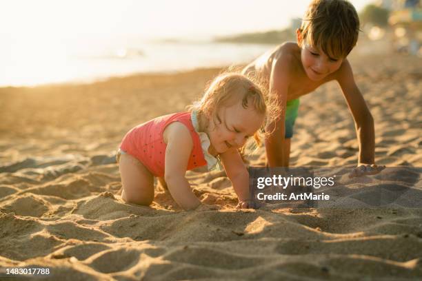 cute happy siblings, baby girl and her brother, play with sand on the beach near the sea on summer holidays in sunset - happy holidays family stock pictures, royalty-free photos & images