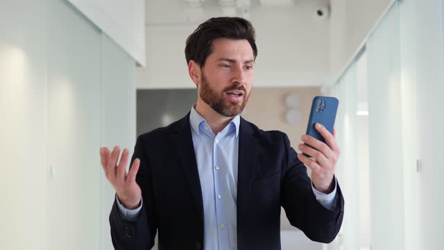 Handsome smart bearded businessman uses his mobile for a professional video call from office communicating with his business partners or colleagues. Man providing updates on current projects.