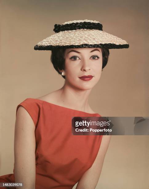 Posed studio portrait of a female fashion model wearing an orange sleeveless boat necked dress and a two tone straw hat, London, 17th May 1958.