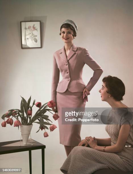 Posed studio portrait of two female fashion models wearing, standing in centre, a lilac tailored skirt suit and, seated on right, a sleeveless...
