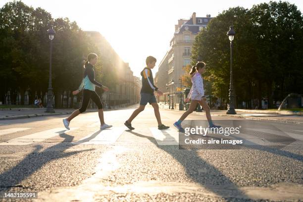 profile view of a family of four wearing casual clothes crossing a street in paris, in the background are trees and a building. - cross road children stock pictures, royalty-free photos & images