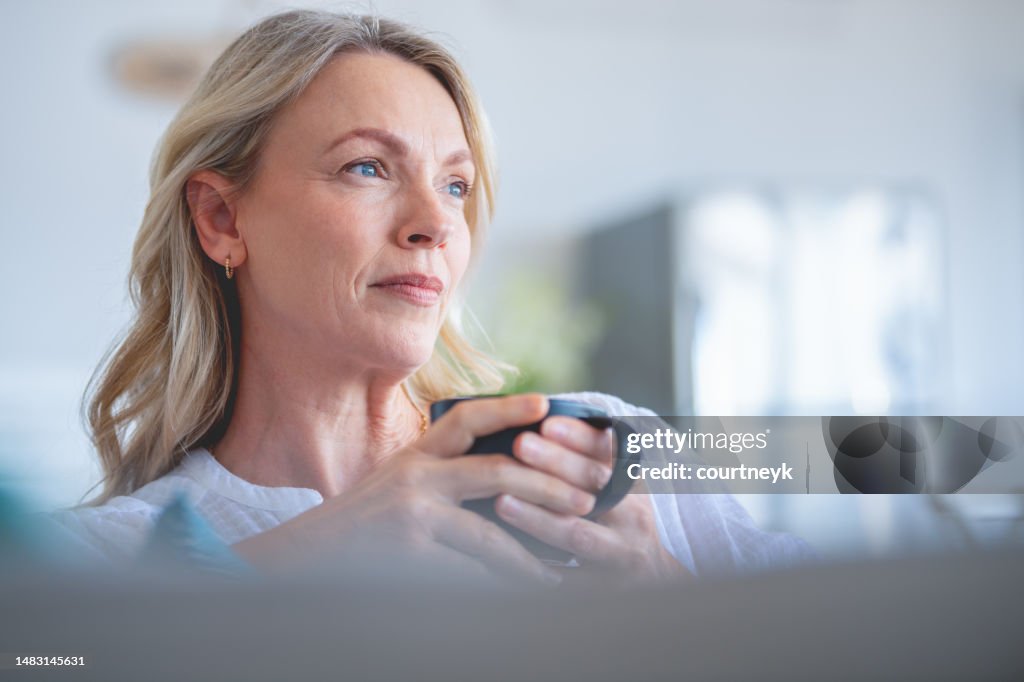 Mature woman drinking a cup of tea or coffee sitting on the sofa.