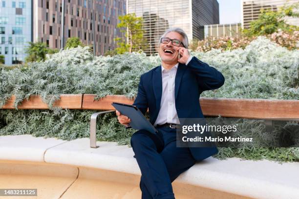 asian businessman laughing and talking on his smartphone while using a digital tablet - man suit using phone tablet foto e immagini stock
