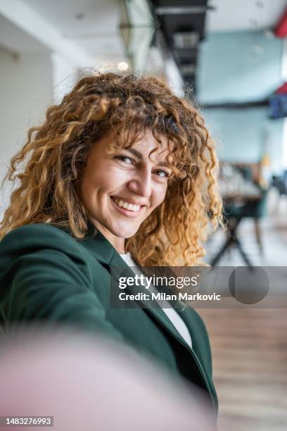 hermosa joven con cabello rizado tomándose selfie mientras está de pie en la cafetería, vestida con blazer verde, casual elegante - mujeres de mediana edad fotografías e imágenes de stock