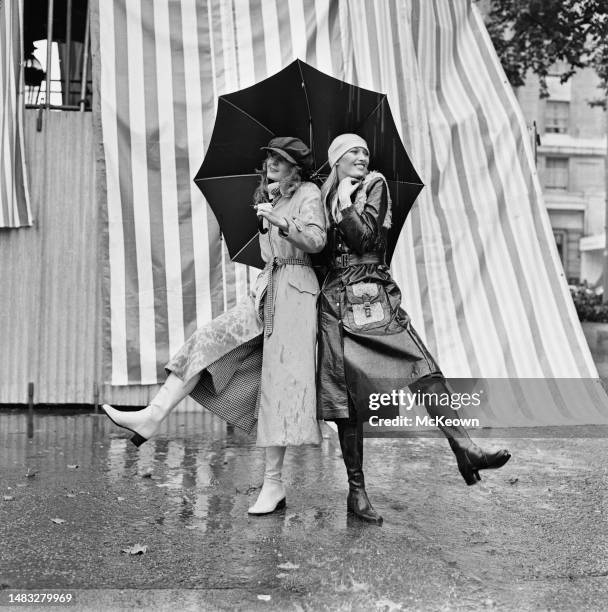 Two female fashion models, one wearing a beige gabardine trench coat, the other wearing a dark brown polyurethane raincoat, both standing beneath a...