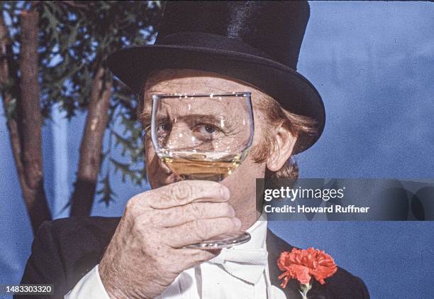 Portrait of American actor John Byner, in formal evening wear and a top hat, as he looks through a wineglass on the set of a corporate video,...