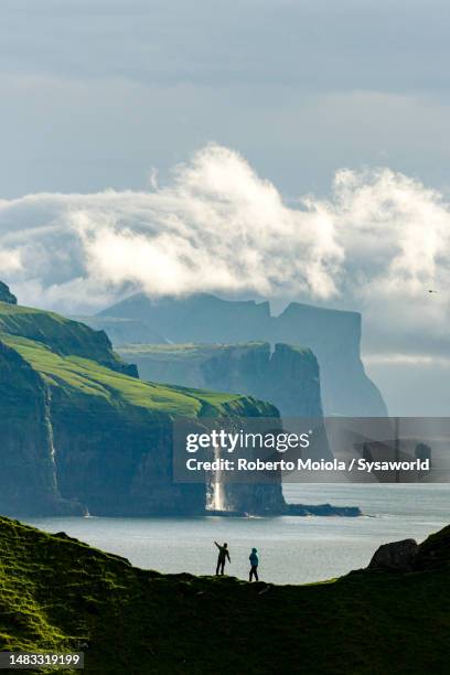 silhouette of tourists admiring a waterfall from cliffs - faroe islands photos et images de collection