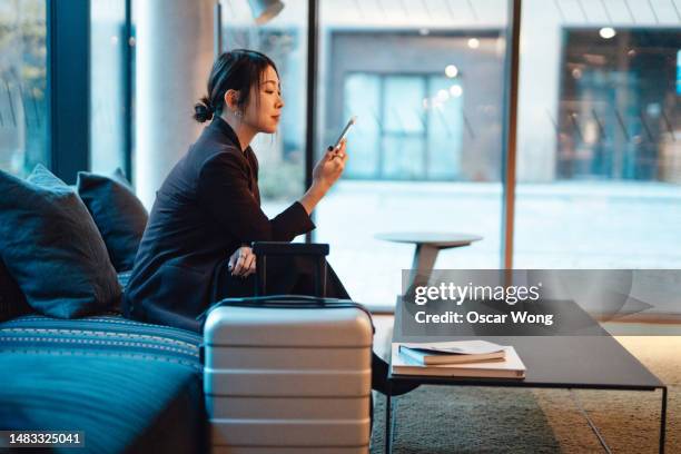 young asian business woman using smartphone sitting at hotel lobby with suitcase - business travel hotel stock pictures, royalty-free photos & images