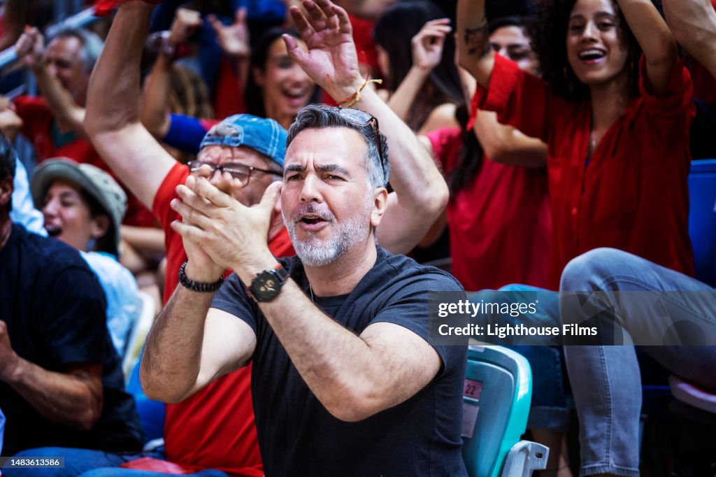 Thrilled soccer fans excitedly clap and cheer sitting next to each other in the stadium bleachers