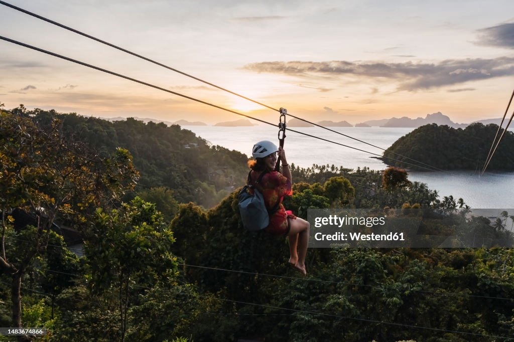 Woman riding on zip line over tree at sunset