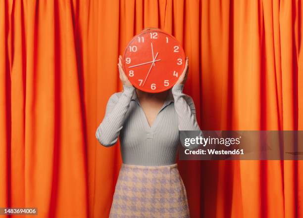 woman holding red clock over face in front of orange curtain - frau uhr stock-fotos und bilder