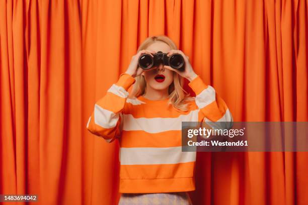 woman looking through binoculars standing in front of orange curtain - canocchiale foto e immagini stock