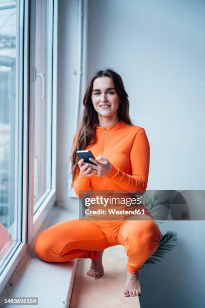 businesswoman crouching with smart phone on window sill in office - jumpsuit stockfoto's en -beelden