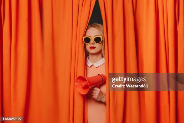 woman wearing sunglasses standing with megaphone amidst curtains - advertisement stock-fotos und bilder