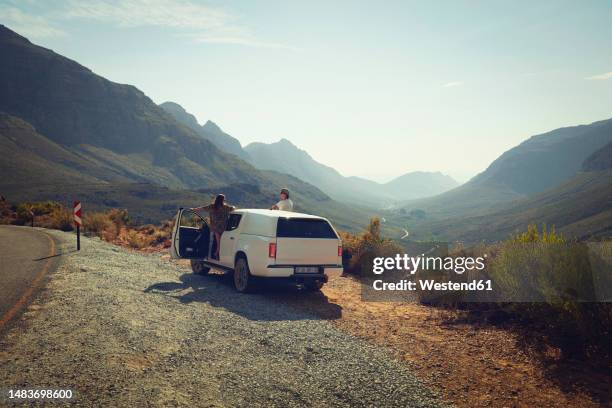 friends watching mountains from car at roadside - borde de la carretera fotografías e imágenes de stock